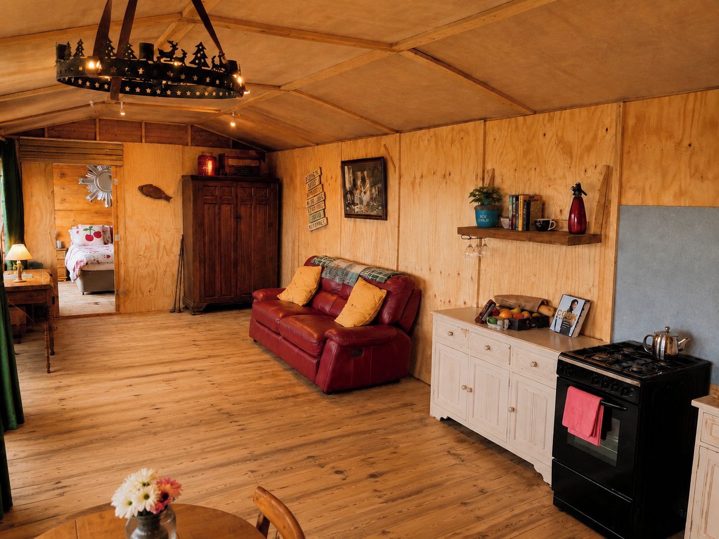 L’intérieur d’un chalet accueillant, avec un plancher en bois, un canapé en cuir rouge, un coin cuisine équipé d’une cuisinière et une table à manger agrémentée d’un vase de fleurs.