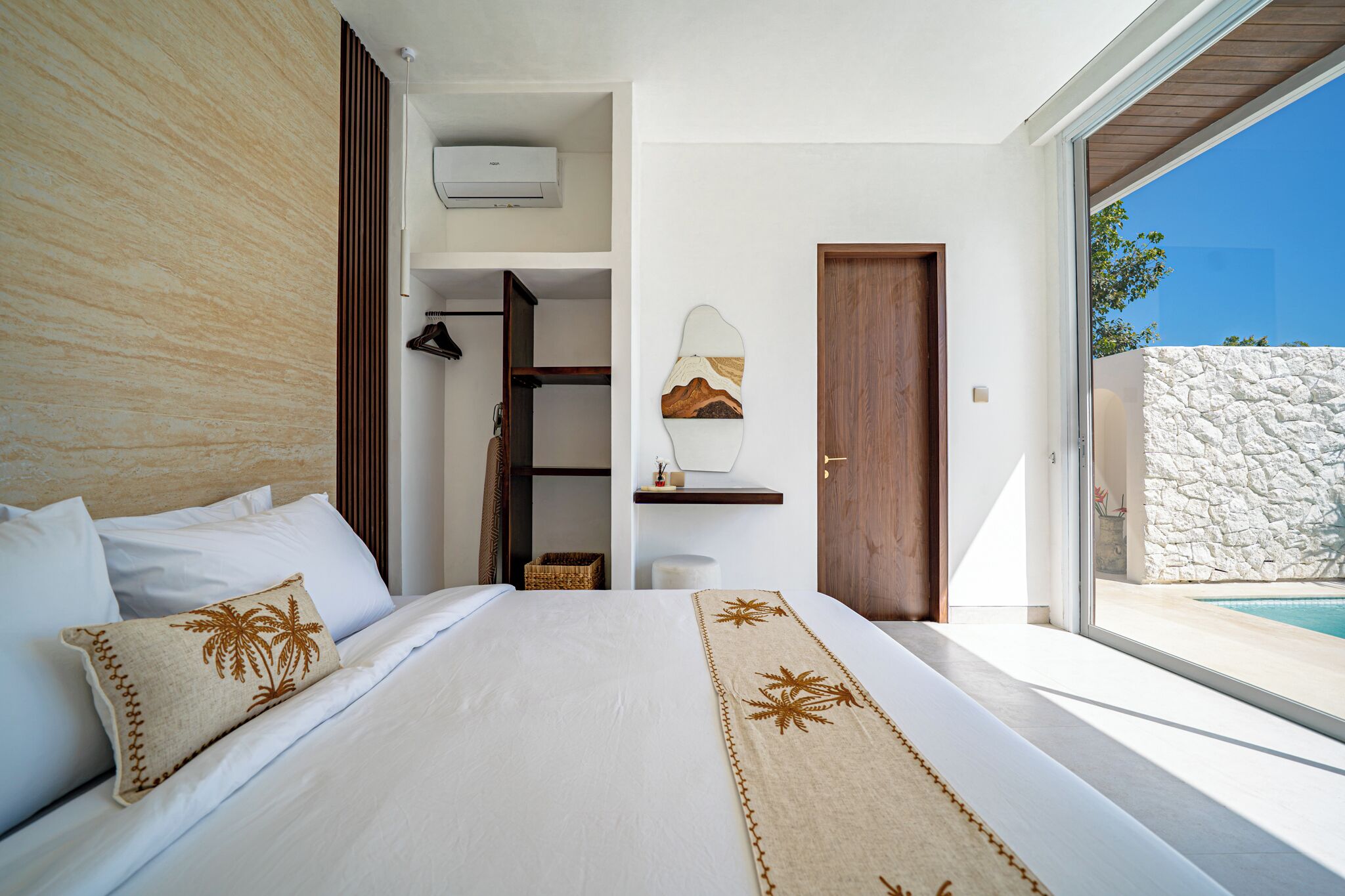 A modern bedroom with a large bed, a built-in wardrobe, a small seating area, and a view of a pool.