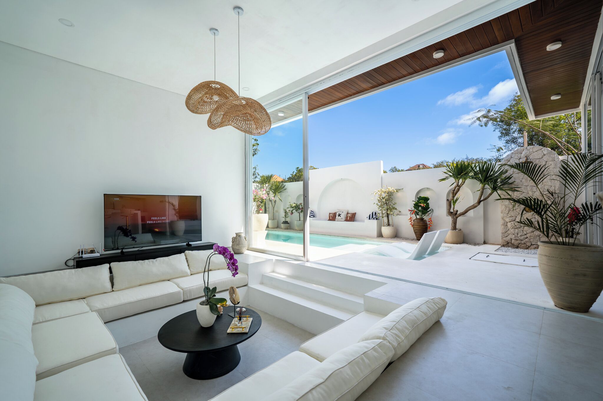 A modern living room with a sofa, coffee table, and TV, overlooking a pool and outdoor area with plants.