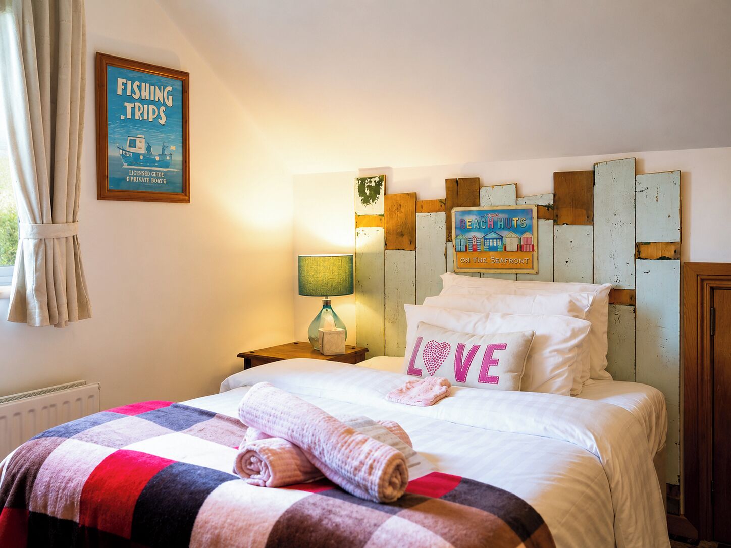 Une chambre d’hôtel avec un lit, une table de chevet, une lampe, un tableau encadré représentant une excursion de pêche et une décoration murale représentant des cabanes de plage.