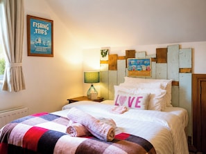 A hotel room with a bed, a nightstand, a lamp, a framed picture of a fishing trip, and a wall decoration of beach huts.