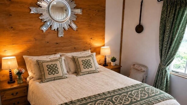 A cozy bedroom with a wooden headboard, a decorative mirror, two bedside lamps, a patterned throw blanket, and a window with curtains. - Cottage Sleeps 4 with Parking and Sea Views (Carrowmenagh)