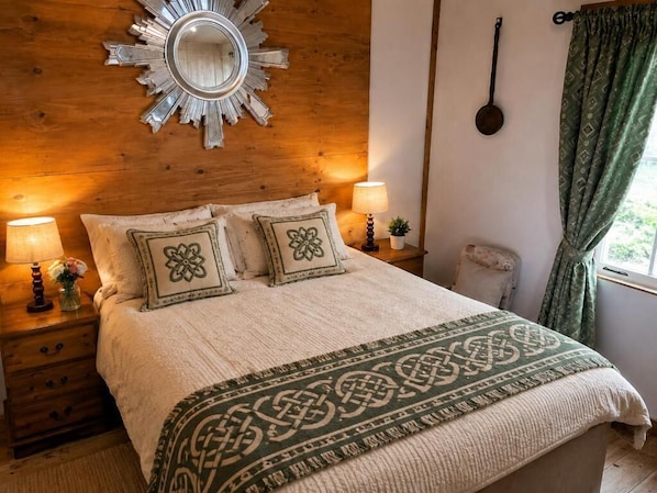 Ein gemütliches Schlafzimmer mit einem Holz-Kopfteil, einem dekorativen Spiegel, zwei Nachttischlampen, einer gemusterten Tagesdecke und einem Fenster mit Vorhängen.