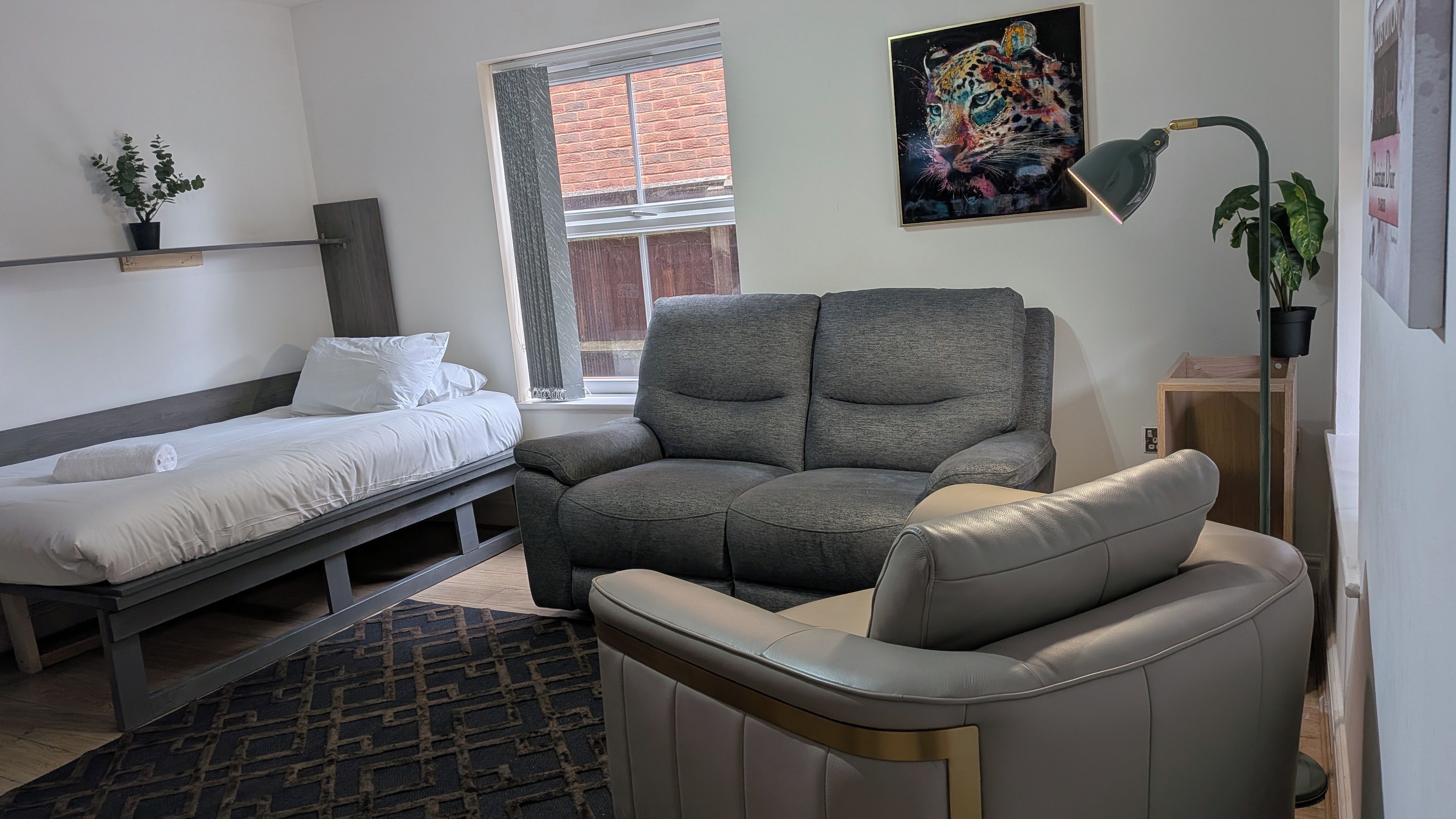 Een moderne slaapkamer met een bed, een grijze bank, een klein tafeltje, een kamerplant en een raam met uitzicht.