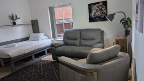 Une chambre moderne avec un lit, un canapé gris, une petite table, une plante en pot et une fenêtre donnant sur un beau paysage.