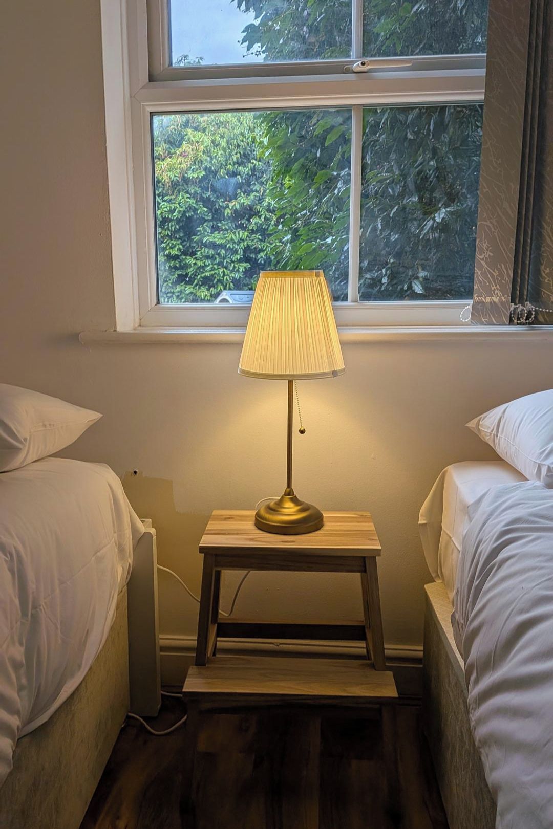 Une chambre avec deux lits, une table de chevet avec une lampe, une fenêtre donnant sur un espace verdoyant et un radiateur.