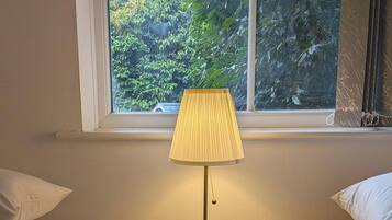 A bedroom with two beds, a bedside table with a lamp, a window with a view of greenery, and a radiator.