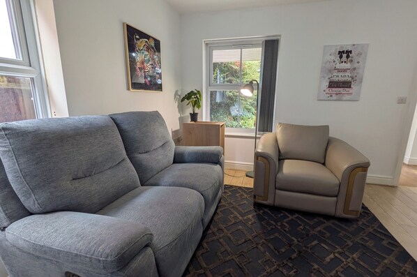 Un salon avec un canapé gris, un fauteuil beige, une petite table d’appoint en bois et une plante en pot.