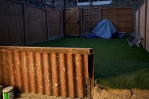 Ein umzäunter Garten mit Rasen aus Kunststoff, einem überdachten Bereich und einer Metallkonstruktion.