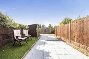 Ein Garten mit einer gepflasterten Terrasse, zwei gestreiften Liegestühlen, einem kleinen Schuppen und einem Holzzaun.