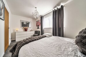 Une chambre à coucher comprenant un lit, une commode, une chaise, un lustre, une fenêtre avec des rideaux et un tableau accroché au mur.