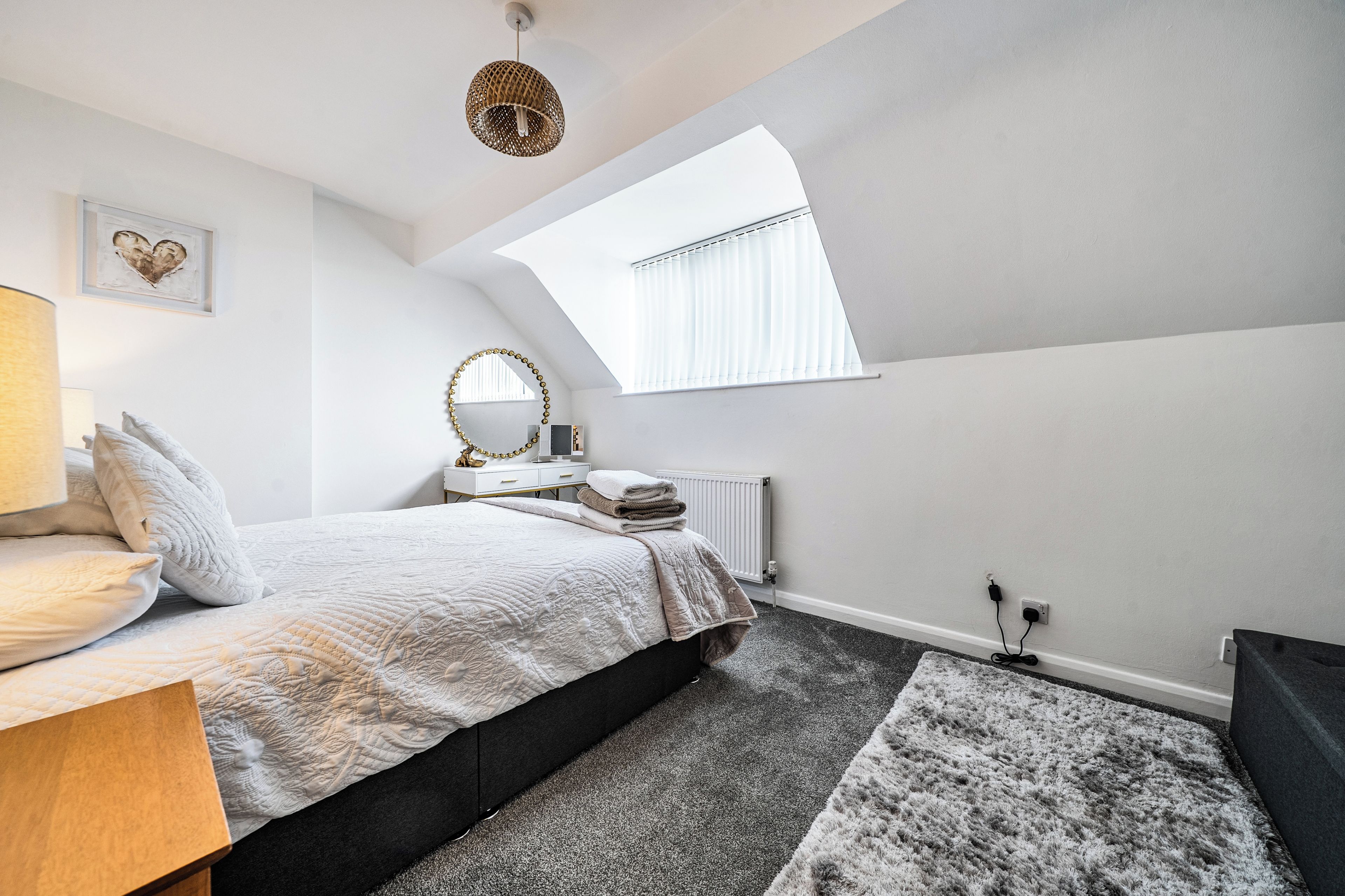 A bedroom with a bed, bedside table, lamp, mirror, radiator, and a window with blinds.