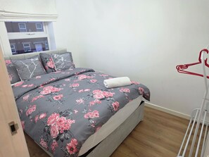 A bedroom with a bed, grey floral bedding, a white towel, and a red hanger rack.