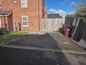 A paved driveway leading to a wooden gate, a brick building with a satellite dish, and a red trash bin.
