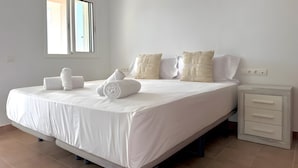 A neatly made bed with white linens and rolled towels, a bedside table with drawers, and a window with blinds. - Can Alaya - Luxury Holiday Villa in Jesús, Ibiza with Views of Dalt Vila (Santa Eulària des Riu)