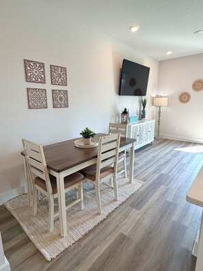 Een eetgedeelte met een houten tafel en stoelen, een geweven vloerkleed en een flatscreen aan de muur.