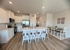 A modern kitchen with white cabinetry, a central island with bar stools, a stainless steel refrigerator, and a dining area with a wooden table and chairs. - Gulf Views + Golf Cart & Pools! (Port St. Joe)