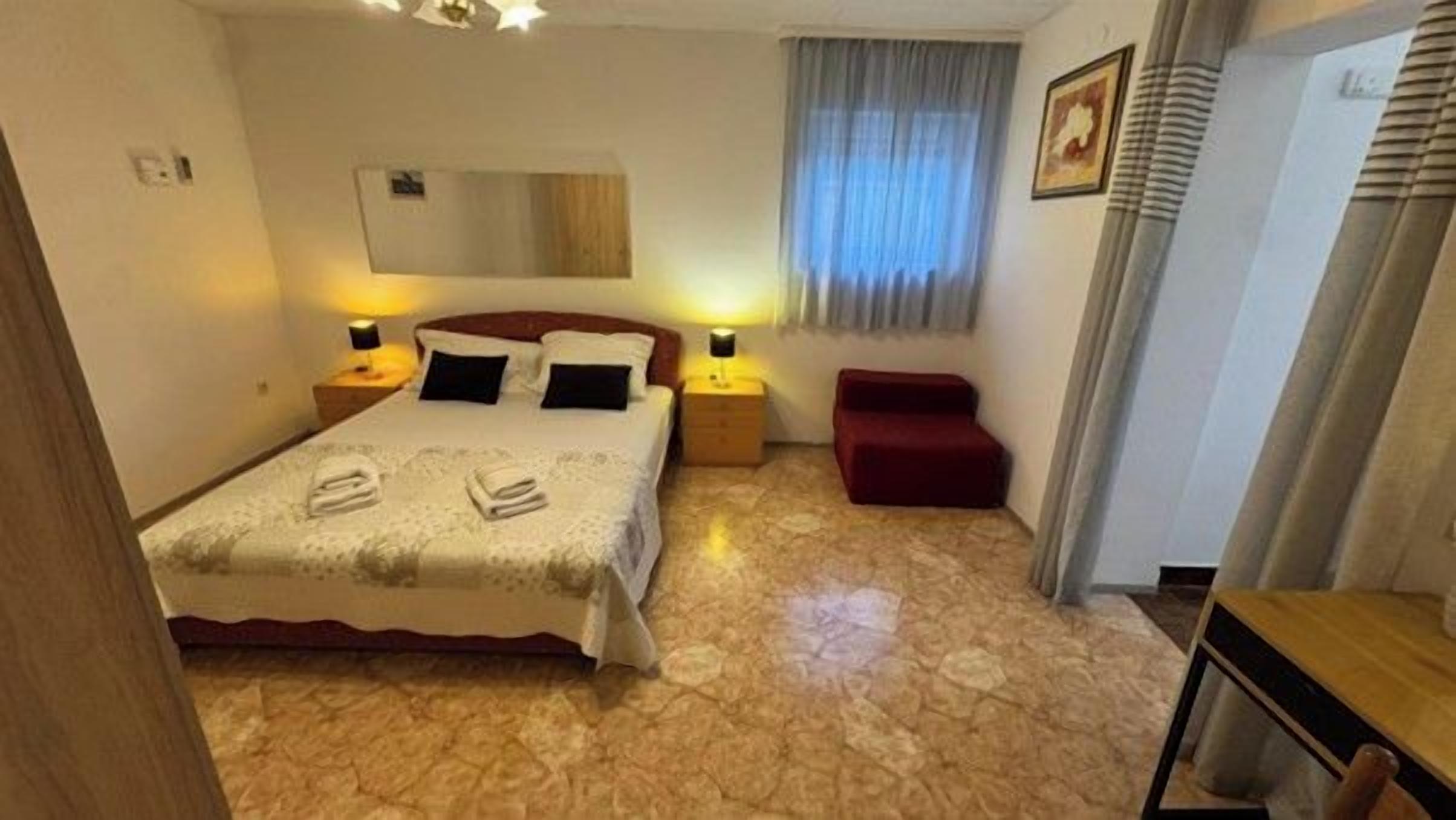 A hotel room with a bed, bedside lamps, a red bench, a wooden table, and a window with curtains.