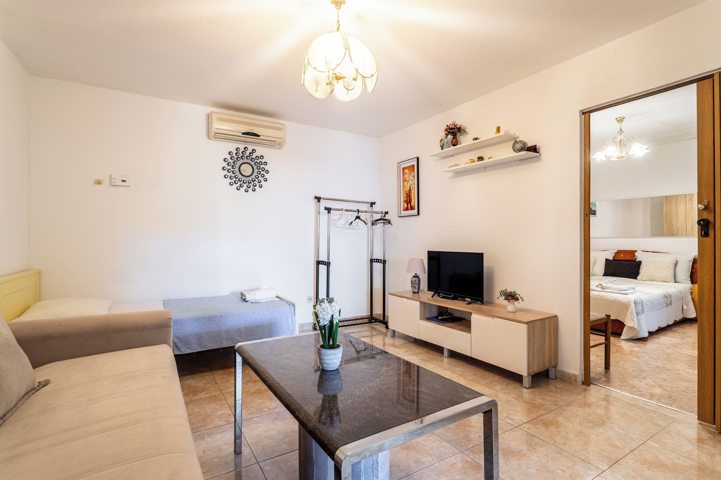Ein modernes Wohnzimmer mit einer Couch, einem Couchtisch, einem Fernseher und einem kleinen Esstisch. Im Hintergrund steht ein Bett und an der Wand hängt ein Spiegel.