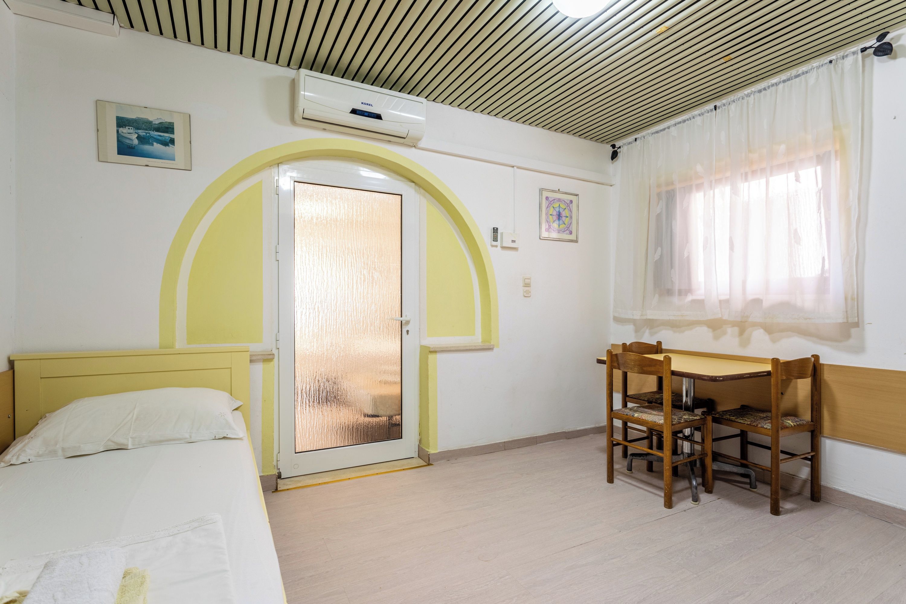 Een kamer met een bed, een tafel met stoelen, een raam met gordijnen, een airconditioning en een deur met ruit.