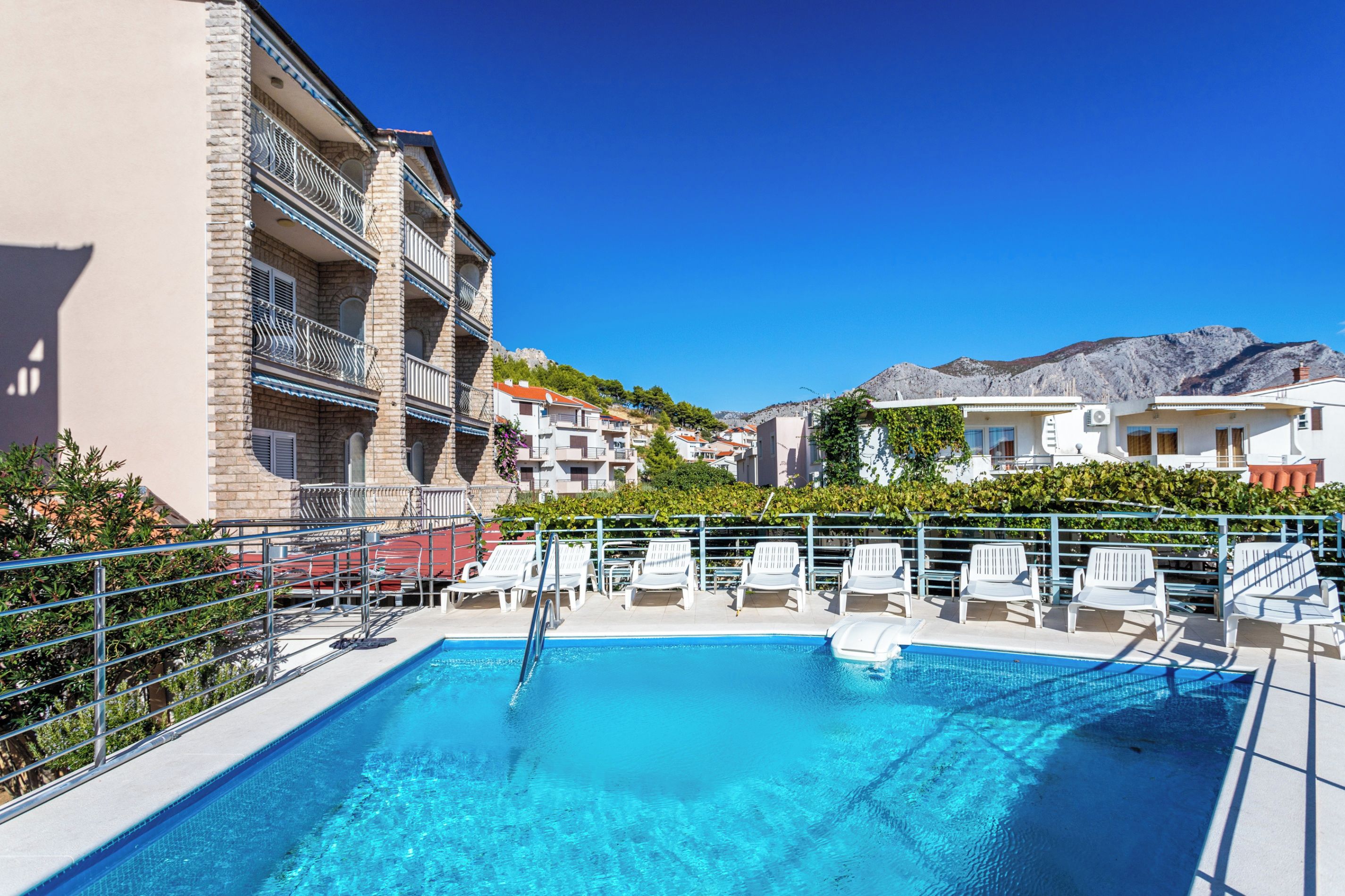 A swimming pool with lounge chairs, a stone building with balconies, and a mountain in the background.