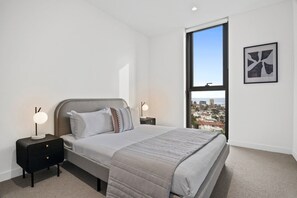 Une chambre à coucher avec un lit, une table de chevet, une lampe, une fenêtre donnant sur la ville et un tableau encadré au mur.