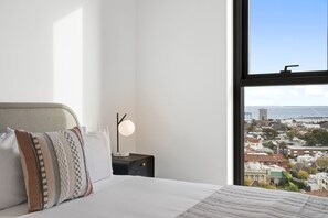 A bedroom with a bed, pillows, a bedside table with a lamp, and a window with a city view. - Balcony 3-Bed Apartment Near Albert Park (St Kilda)