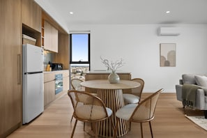A modern kitchen with a dining area, featuring a round table with chairs, a view through the window, and a living area with a sofa. - Sky-View 2-Bed Apartment Near St Kilda Beach (St Kilda)