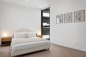 A bedroom with a bed, bedside lamps, a window, and three framed pictures on the wall. - Skydeck 2-Bed Residence Near Albert Park (St Kilda)