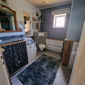 Casa de banho com lavatório, sanita e espelho. Tem uma janela com cortina e um tapete estampado no chão.