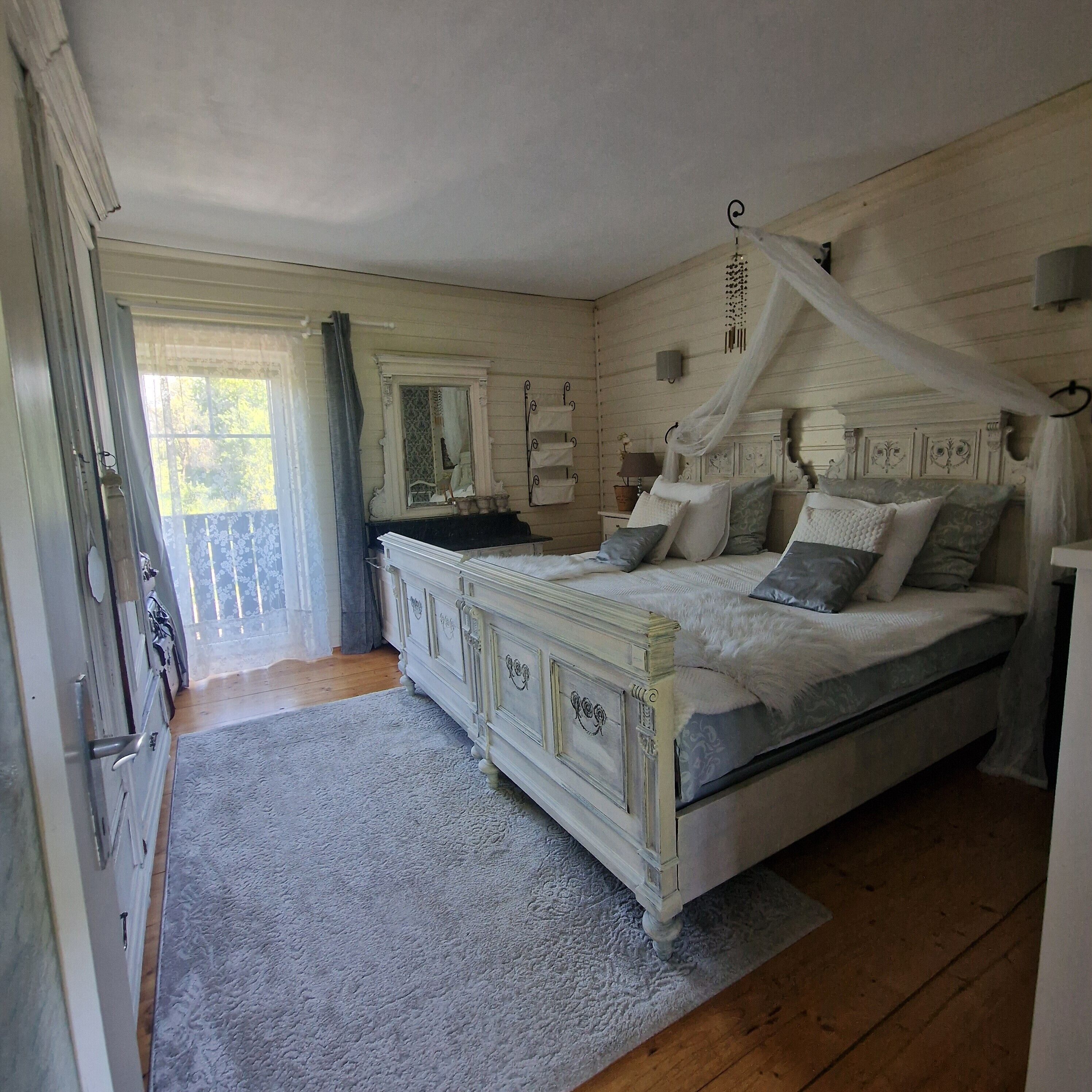 A bedroom with a four-poster bed, a large window with curtains, and a wooden floor.