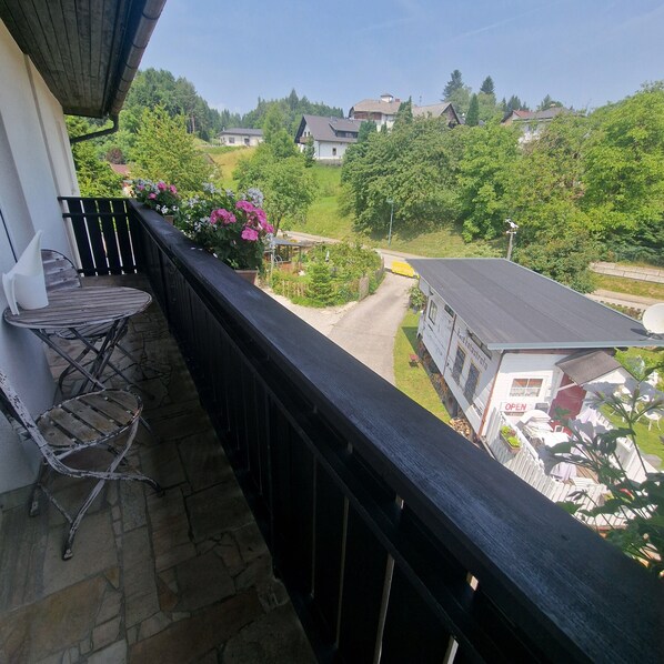 A balcony with a chair, a flower box with pink flowers, and a view of a road and houses in the distance.