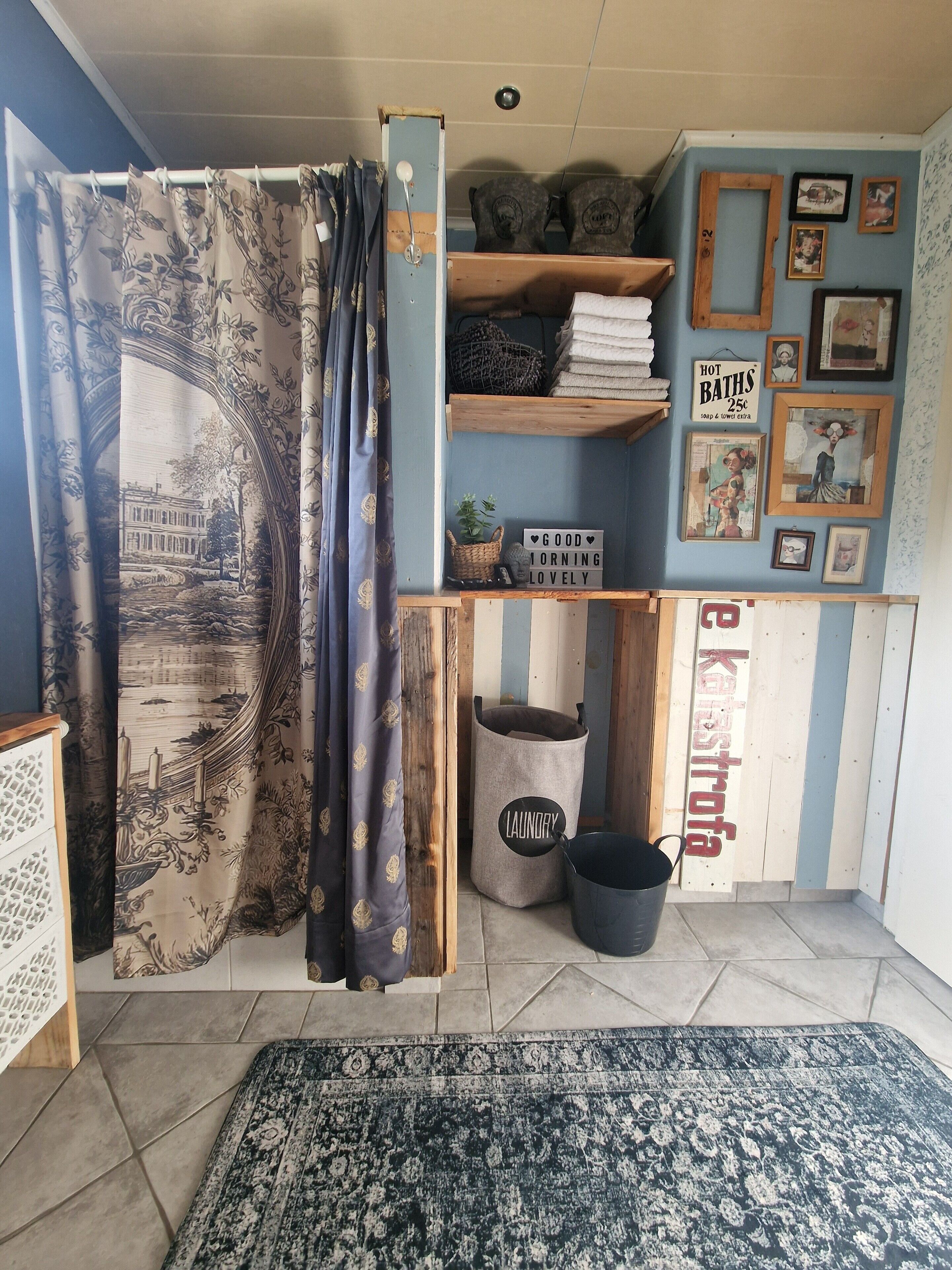 Une pièce avec un rideau représentant un bâtiment historique, une étagère avec des serviettes et un panneau, un panier à linge et un tapis.