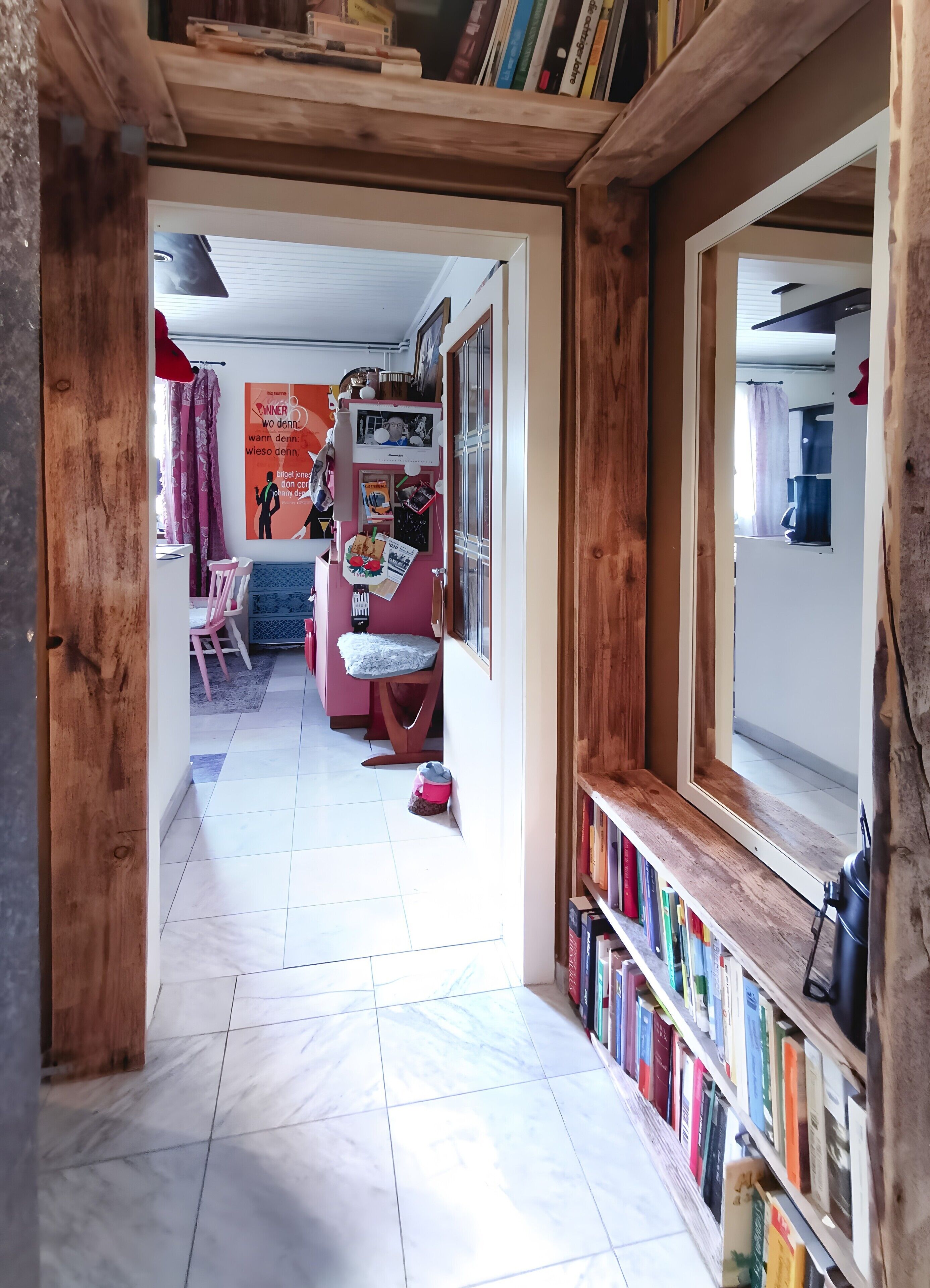 A hallway with a wooden bookshelf, a dining area with a pink chair, and a tiled floor.