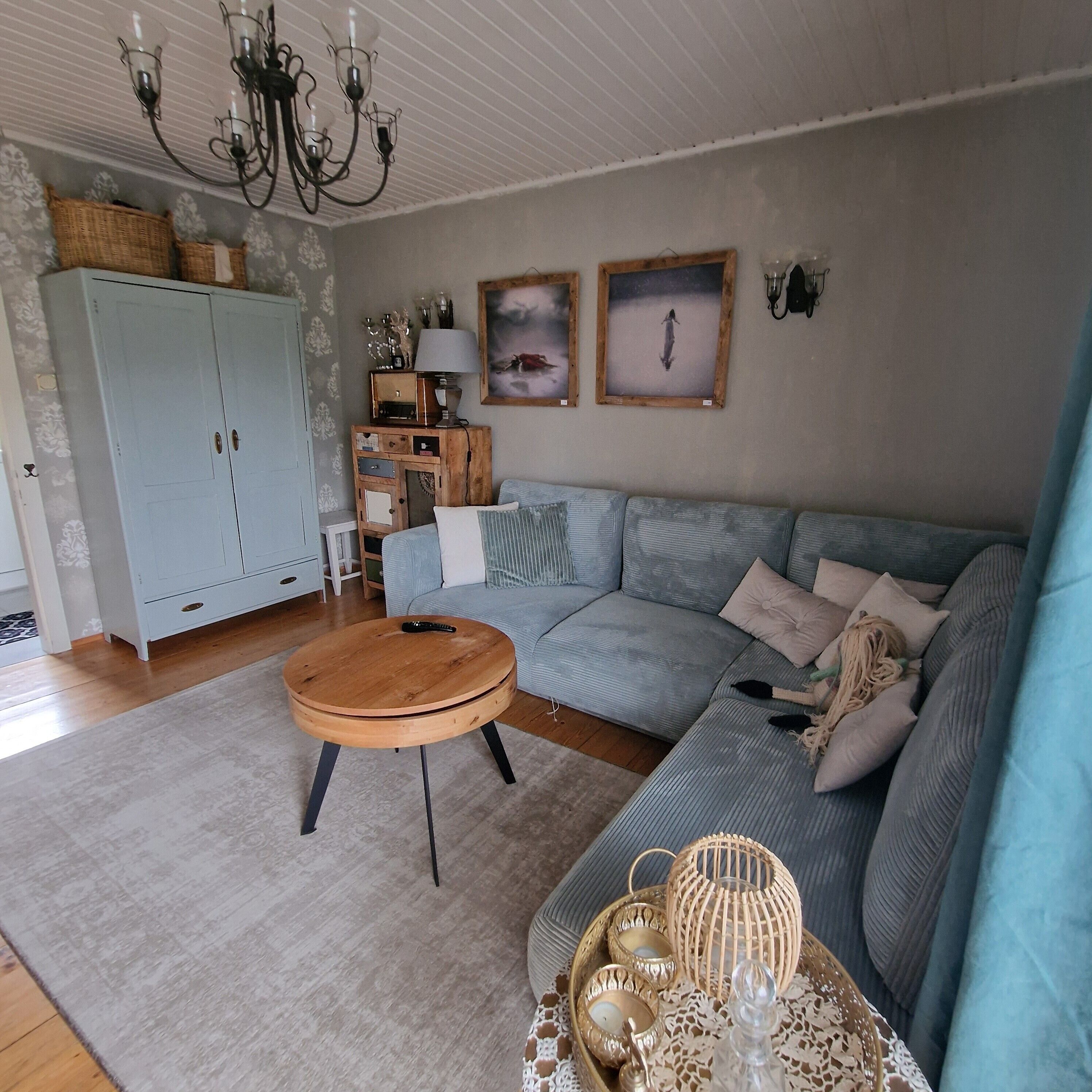 Ein Wohnzimmer mit einem hellblauen Sofa, einem runden Holztisch, einem blau gemusterten Teppich, einer Holzkommode und einer Pendelleuchte.