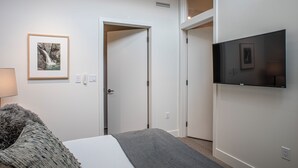 Un dormitorio con cama, televisor de pantalla plana montado en la pared, puerta que da a otra habitación y un cuadro enmarcado.