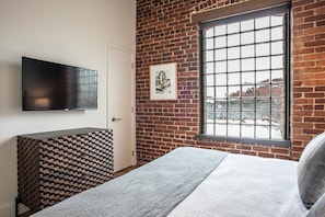 Une chambre avec un mur en briques, une fenêtre avec des barreaux en métal, un téléviseur à écran plat, un tableau encadré et une commode à motifs.