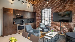 Una cocina moderna con electrodomésticos integrados, un comedor con mesa y sillas, y una sala de estar con sofá y televisor montado en una pared de ladrillo.