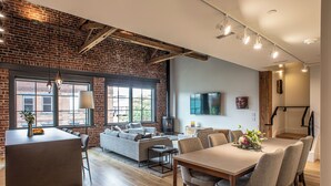 A modern living space with a brick wall, large windows, a fireplace, and a dining area. - Elevation Lofts Hotel - Broadway Loft Collection - Up to 8 Guests (Asheville)