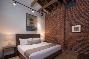 Quarto com parede de tijolos, cama com roupa de cama branca, mesa de cabeceira com abajur e um quadro na parede.