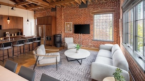 A modern kitchen with a brick wall, a flat-screen TV, and a dining area with a round coffee table. - Elevation Lofts Hotel - Group Loft Collection - Up to 14 Guests (Asheville)