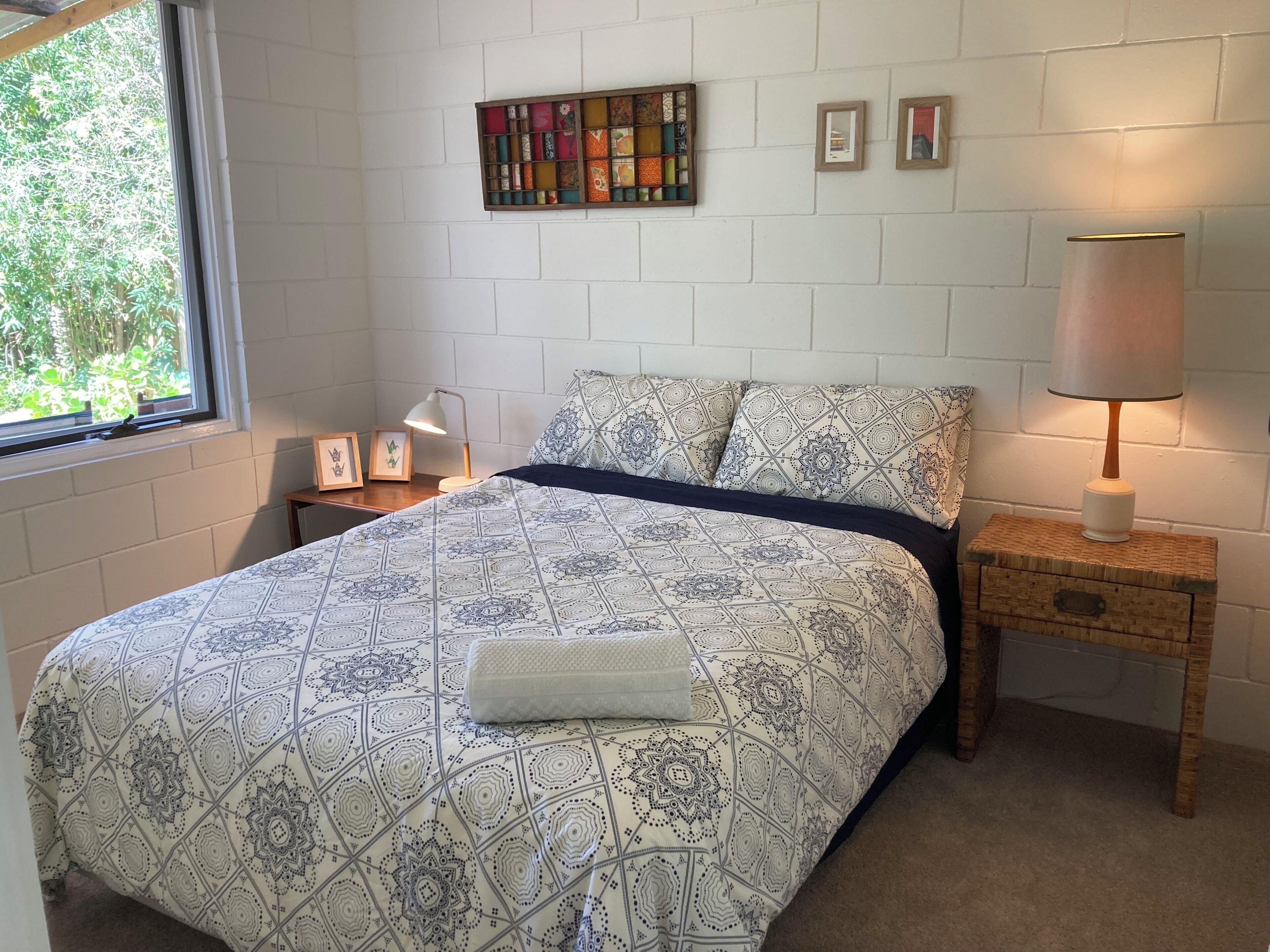 A bedroom with a patterned bedspread, a bedside lamp, a small wooden nightstand, and a wall-mounted shelf with decorative items.