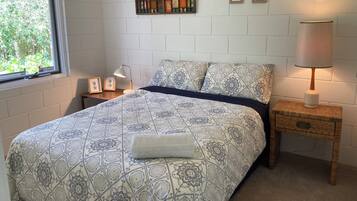 A bedroom with a patterned bedspread, a bedside lamp, a small wooden nightstand, and a wall-mounted shelf with decorative items.