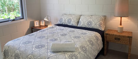A bedroom with a patterned bedspread, a bedside lamp, a small wooden nightstand, and a wall-mounted shelf with decorative items.
