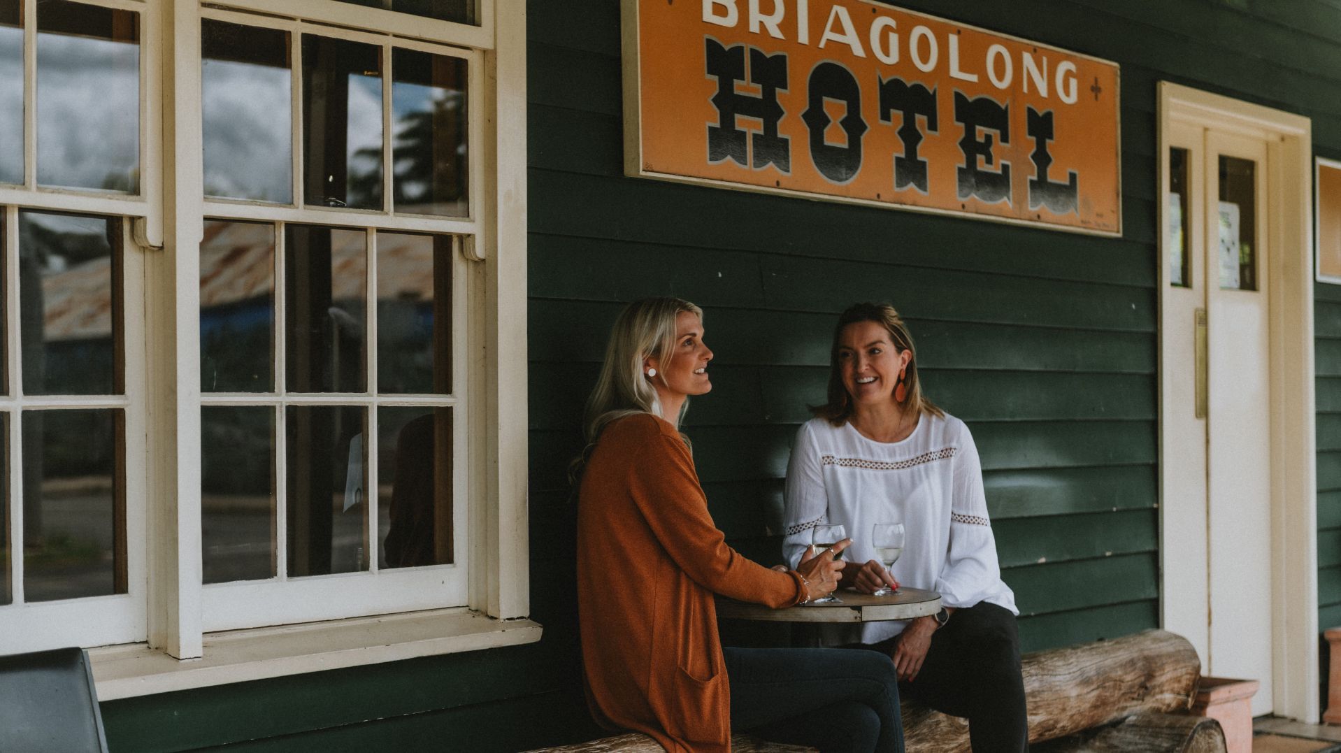 Zwei Personen sitzen an einem Tisch vor einem Gebäude, an dem ein Schild mit der Aufschrift 'BRIAGOLONG HOTEL' hängt.