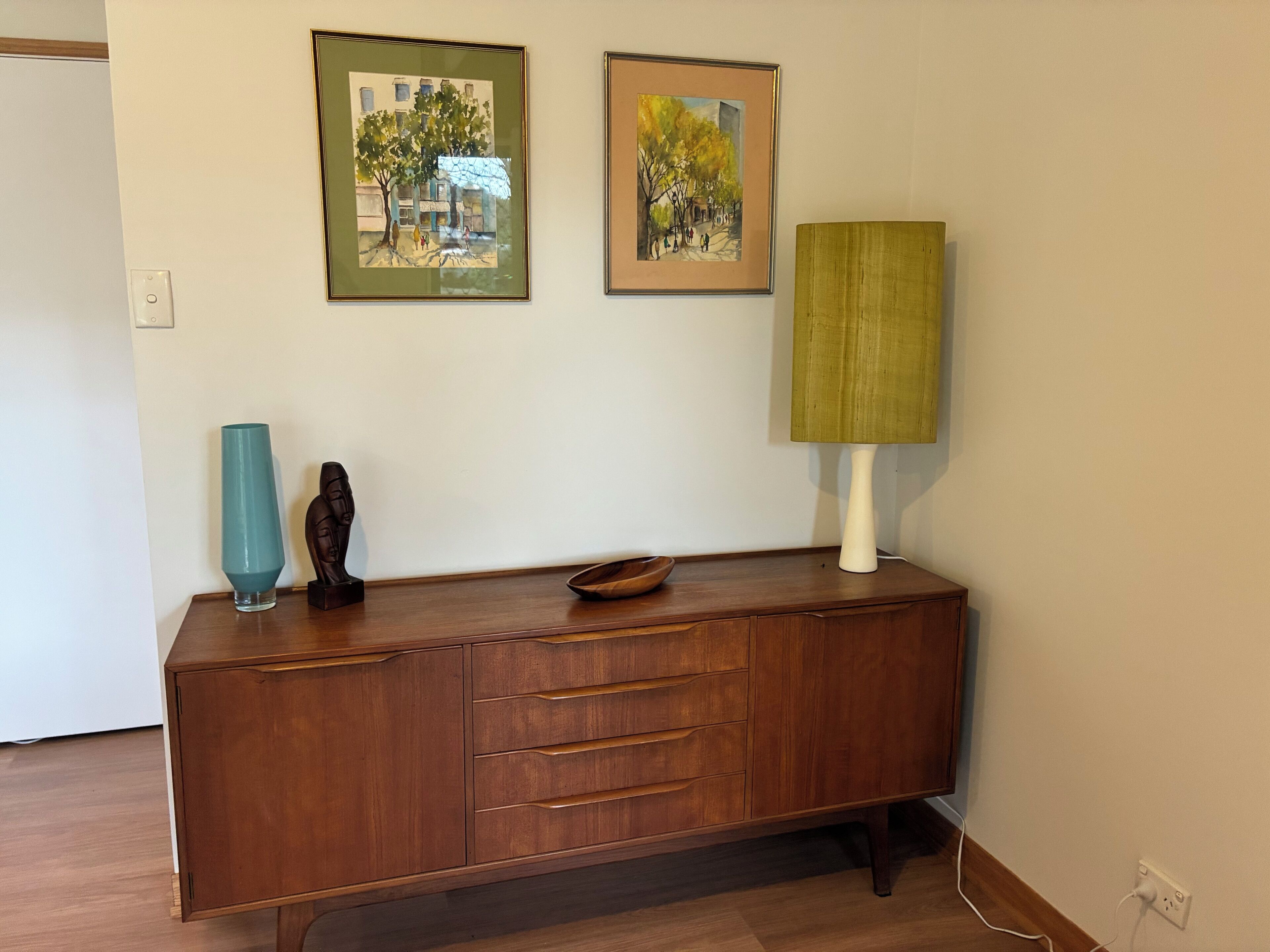 A wooden cabinet with drawers and a vase, two framed paintings, and a lamp on a table.