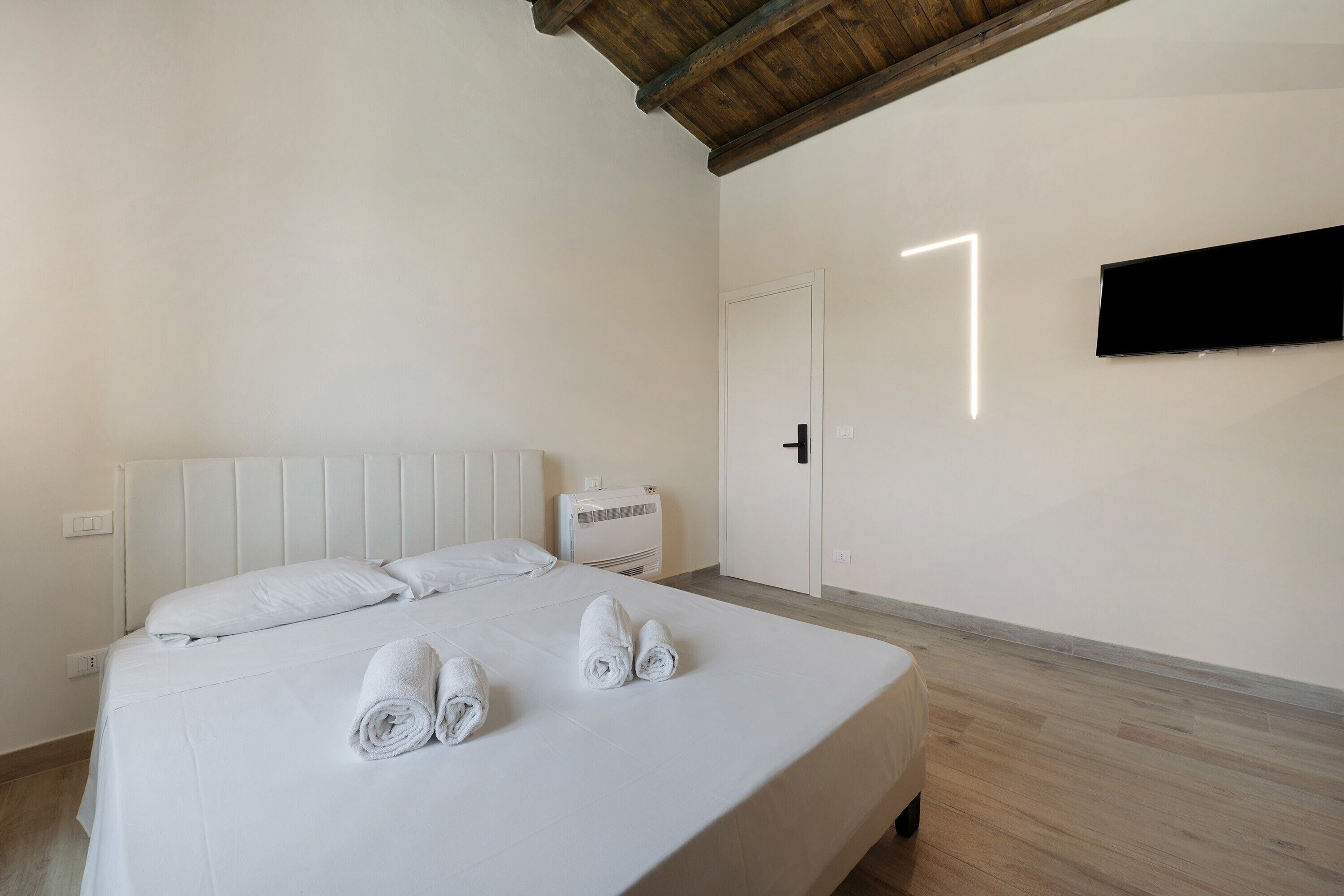 A hotel room with a bed, a flat-screen TV mounted on the wall, and a door with a light switch.