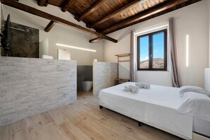 A bedroom with a wooden ceiling, a large window with a view, a bed with white linens, and a bathroom with a modern design. - Micu Room - Sikania Agrirelais, Mountain View (Caccamo)