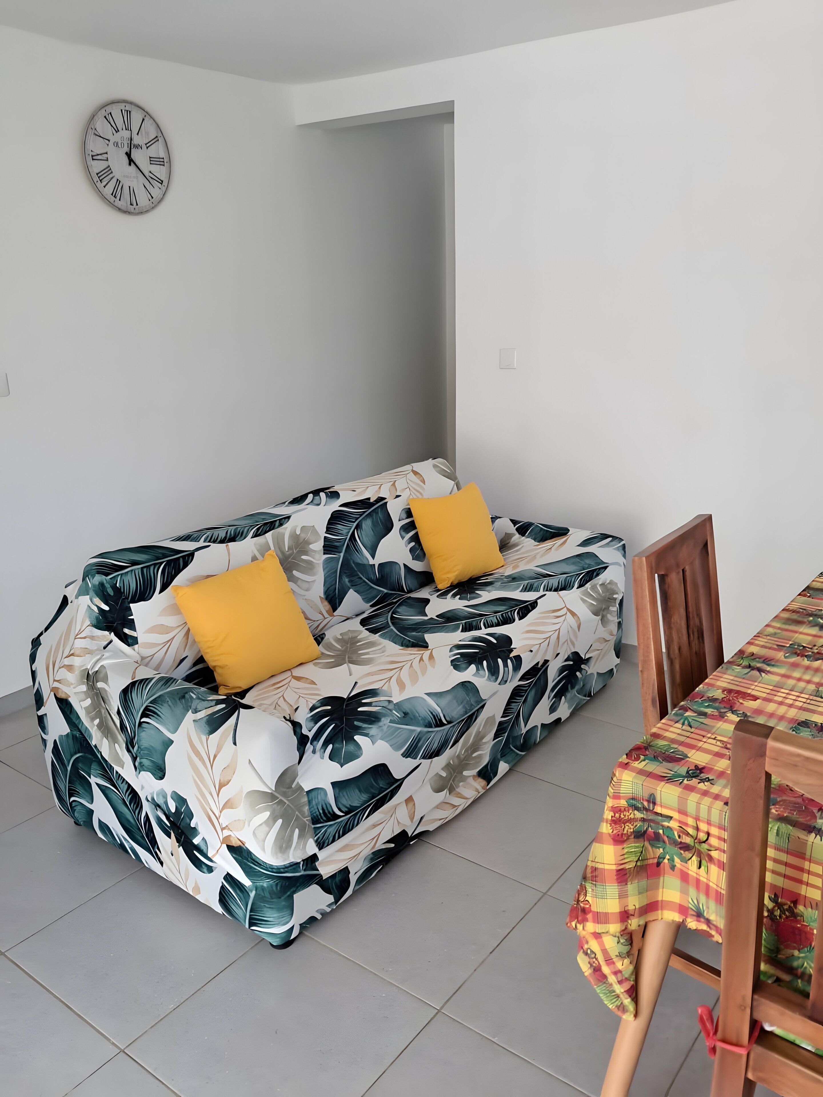A room with a sofa bed featuring a tropical leaf pattern, a wooden dining table with a checkered tablecloth, and a wall clock.