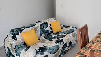 A room with a sofa bed featuring a tropical leaf pattern, a wooden dining table with a checkered tablecloth, and a wall clock.
